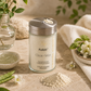 A glass jar of PuRéfi Clear-Skin Exfoliating Cleanser for Acne-Prone skin with a silver lid, styled on a beige stone surface with a small pile of the cream-colored powder, white flowers, and green leaves.