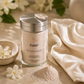 A glass jar of PuRifi Immortal Luxury Organic Anti-Aging Exfoliating Scrub with a silver lid, styled on a beige surface with a small pile of the pinkish-beige scrub, white flowers, and a cream-colored towel.