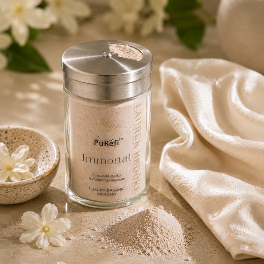 A glass jar of PuRifi Immortal Luxury Organic Anti-Aging Exfoliating Scrub with a silver lid, styled on a beige surface with a small pile of the pinkish-beige scrub, white flowers, and a cream-colored towel.