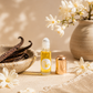 A small glass roller bottle filled with golden yellow botanical vanilla fragrance oil stands on a beige fabric surface beside a metallic rose-gold cap. The bottle has a white label with a yellow Lauren Brooke leaf logo.